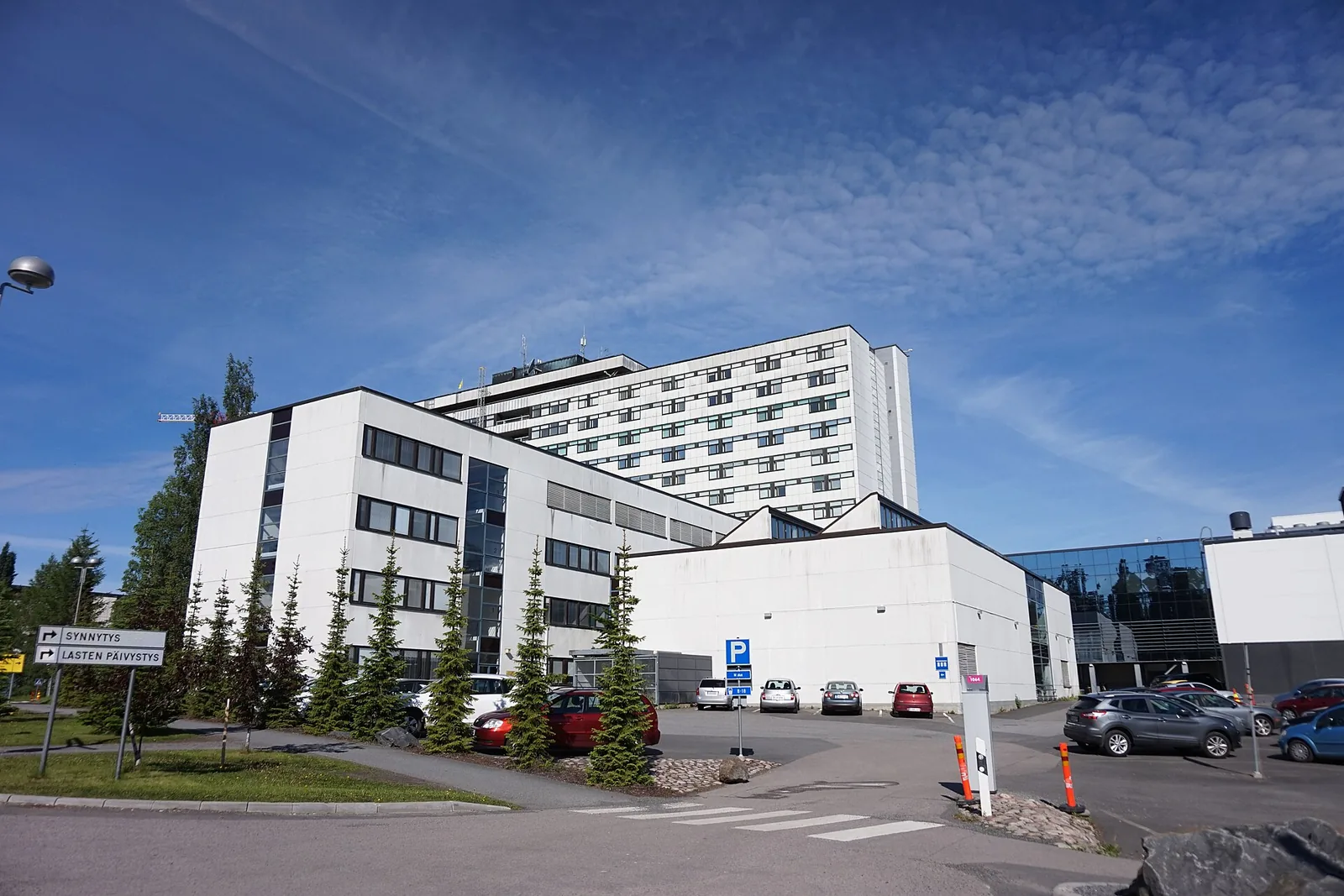 Suuri valkoinen sairaalarakennus ja pysäköintialue sinistä taivasta vasten