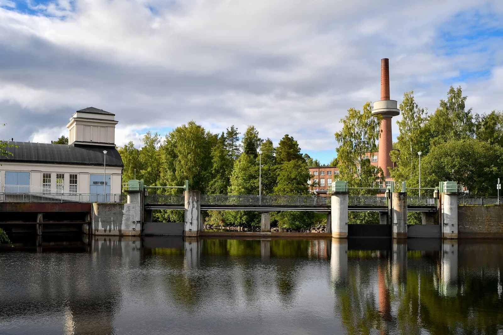Historiallinen pato ja vesilaitos kesäisessä maisemassa, jonka taustalla näkyy tiilinen savupiippu