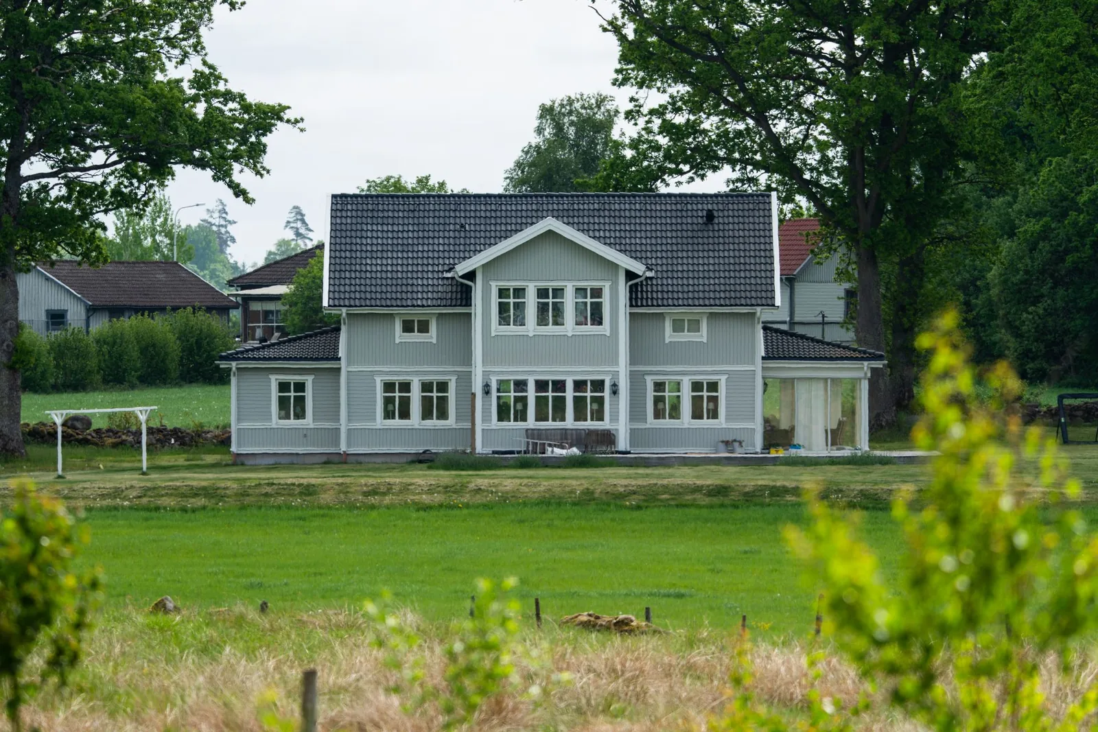 Harmaa kaksikerroksinen omakotitalo niityn ja puiden keskellä